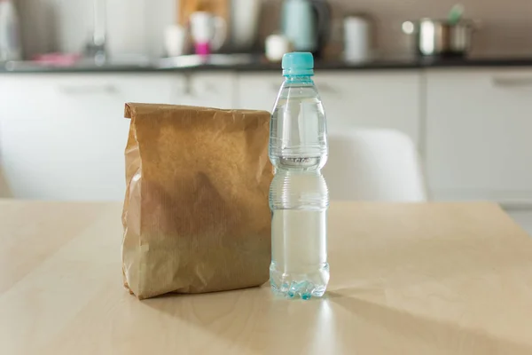 Бумажный пакет для ланча и бутылка воды на деревянном столе на кухонном фоне . — стоковое фото