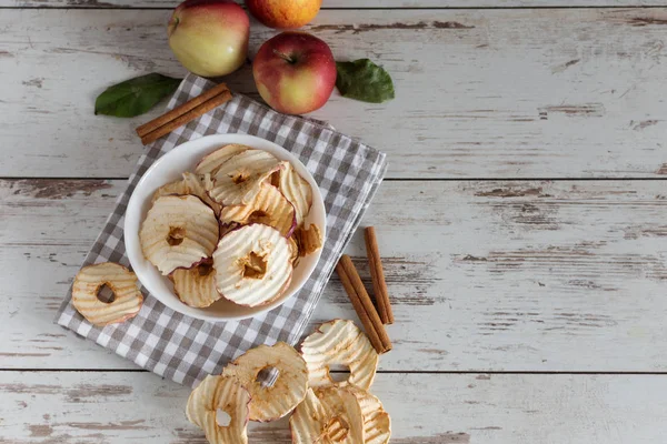 Gedroogde appels chips in schaal met verse rode appels en kaneelstokjes op tafel. — Stockfoto