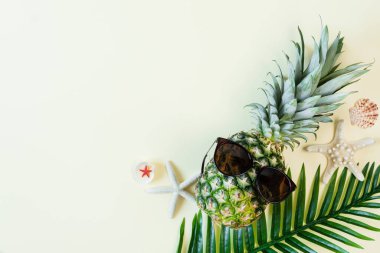 Güneş gözlüklü komik ananas, deniz kabuğu ve sarı pastel arka plan manzaralı palmiye yaprağı. Yaz dönemi. Yaz tatilleri.
