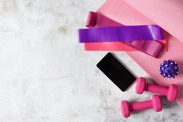 Dumbbells, fitness rubber bands, yoga mat, massage ball and smartphone on white concrete floor. Fitness equipment flat lay. Online workout concept.