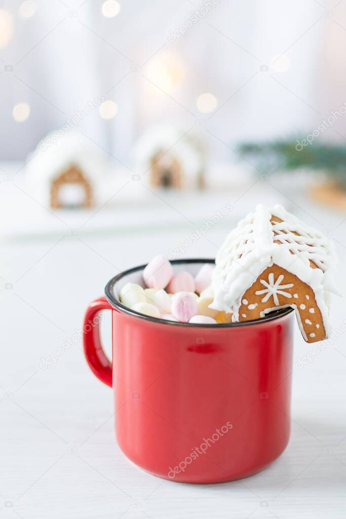 Red mug of hot chocolate with marshmallow and homemade gingerbread small house for decoration on white background in Christmas interior. Christmas holiday drink.