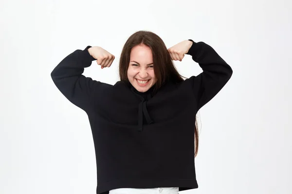 Estudio de una hermosa joven morena con una sudadera negra de pie en doble bíceps posan sobre fondo blanco . — Foto de Stock