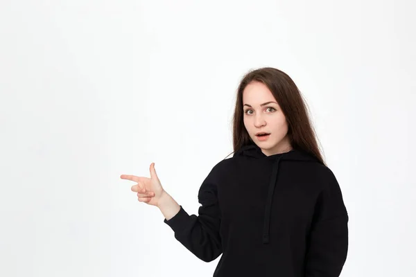 Retrato de una guapa morena joven con una sudadera negra sobre un fondo blanco apuntando con los dedos hacia la izquierda . — Foto de Stock