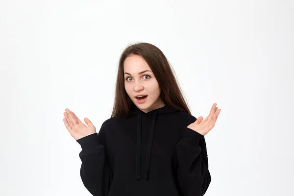 Beyaz arka plan üzerinde duran ve omuzlarını silkme sırasında sürpriz duygularını ifade siyah bir sweatshirt güzel bir genç kadın stüdyo çekim.
