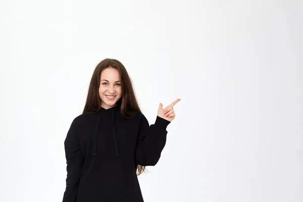 Portrait d'une jolie jeune brune en sweat-shirt noir sur fond blanc pointant le côté droit avec les doigts et regardant la caméra . — Photo