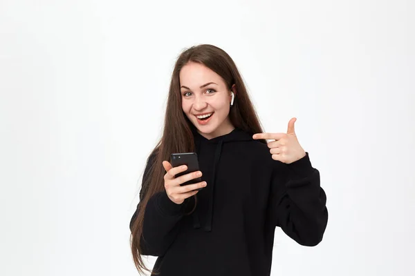 工作室拍摄的一个美丽的年轻黑发女人与无线耳机和手机看着快乐的相机，并指着手机屏幕，而站在白色背景. — 图库照片