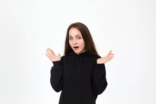 白い背景の上に立ち、肩をすくめながら驚きの感情を表現する黒いスウェットシャツを着た美しい若い女性のスタジオショット. — ストック写真