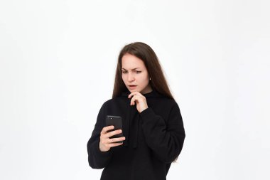 Kablosuz kulaklık ile güzel bir genç esmer kadın stüdyo çekim beyaz arka plan üzerinde dururken telefon şok görünümlü. Kız siyah kapüşonlu giyer.