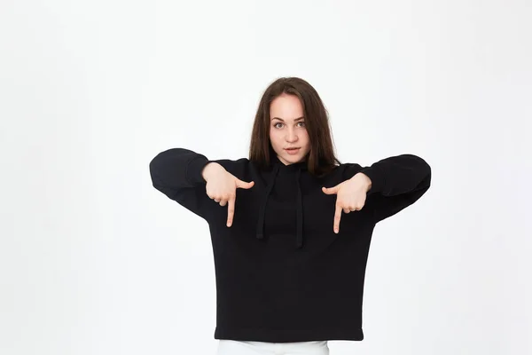 Retrato de una guapa morena joven con una sudadera negra sobre un fondo blanco apuntando hacia abajo con los dedos . —  Fotos de Stock