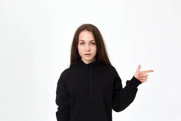 Retrato de una guapa morena joven con una sudadera negra sobre un fondo blanco apuntando al lado derecho con los dedos y mirando a la cámara . —  Fotos de Stock