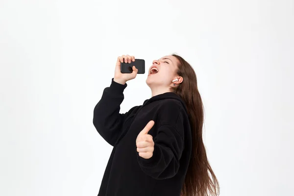 Estúdio de uma bela jovem morena com fone de ouvido sem fio cantando no telefone como se fosse um microfone enquanto estava em pé sobre fundo branco . — Fotografia de Stock