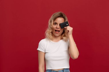 Kırmızı bir arka plan üzerinde duran beyaz bir t-shirt kıvırcık sarı saçlı bir kızın portresi. Model kameraya gülümsüyor ve bir yüzün yarısını kapsayan banka kartı tutar.