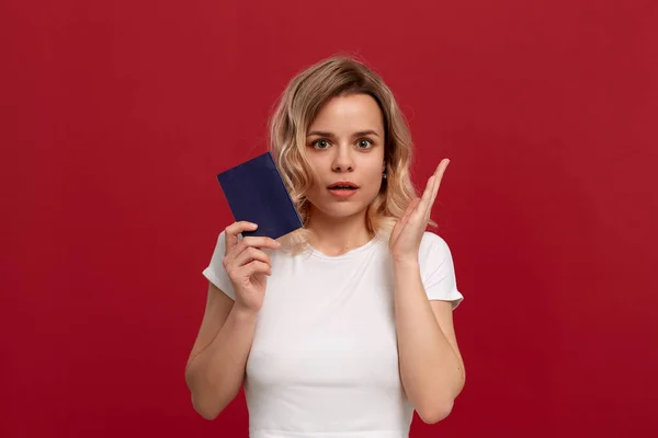 Πορτραίτο ενός όμορφου κοριτσιού με σγουρά ξανθά μαλλιά, ντυμένο σε ένα λευκό μπλουζάκι που στέκεται σε κόκκινο φόντο. Το μοντέλο χαμογελά στην κάμερα, κρατά το διαβατήριο του μπλε χρώματος και ανυψώνει τα χέρια με μια χειρονομία s — Φωτογραφία Αρχείου