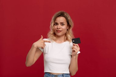 Kırmızı bir arka plan üzerinde duran beyaz bir t-shirt giymiş kıvırcık sarı saçlı güzel bir kız portresi. Model parmak ile banka kartı ve puan tutar 