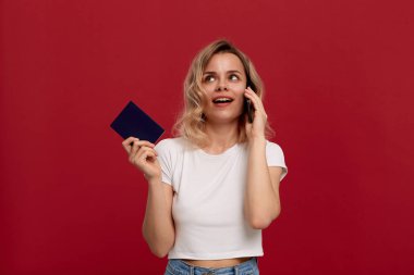 Kırmızı arka plan üzerinde beyaz bir t-shirt kıvırcık sarı saçlı bir kızın portresi. Mutlu model pasaport tutan telefonda konuşuyor.