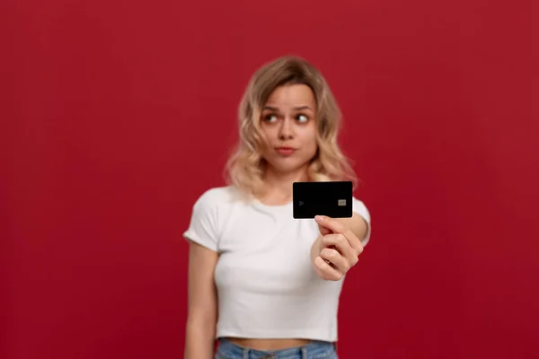 Portrait d'une fille aux cheveux blonds bouclés dans un t-shirt blanc debout sur un fond rouge. Modèle avec une émotion surprise tient la carte bancaire de couleur noire et regarde ailleurs . — Photo