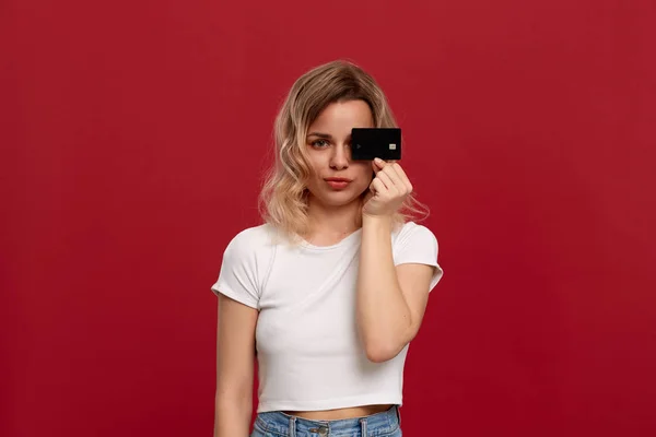 赤い背景に立つ白いTシャツを着た巻き毛のブロンドの髪の少女の肖像画。モデルはカメラを見て、顔の半分を覆う銀行カードを保持します. — ストック写真