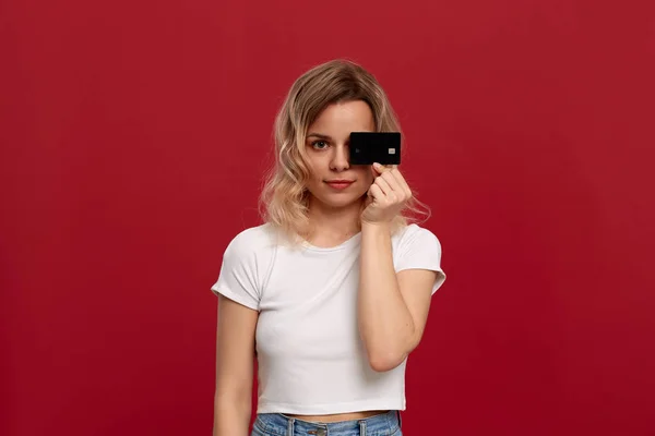 Πορτρέτο ενός κοριτσιού με σγουρά ξανθά μαλλιά σε ένα λευκό μπλουζάκι που στέκεται σε κόκκινο φόντο. Μοντέλο κοιτάζει την κάμερα και κρατά τραπεζική κάρτα που καλύπτει το μισό του προσώπου. — Φωτογραφία Αρχείου