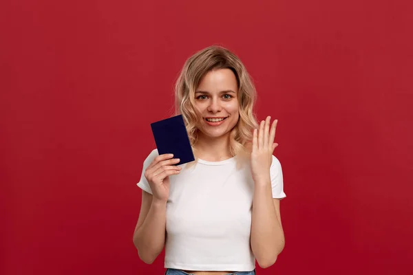 Πορτραίτο ενός όμορφου κοριτσιού με σγουρά ξανθά μαλλιά, ντυμένο σε ένα λευκό μπλουζάκι που στέκεται σε κόκκινο φόντο. Το μοντέλο χαμογελά στην κάμερα, κρατά το διαβατήριο του μπλε χρώματος και ανυψώνει τα χέρια με μια χειρονομία s — Φωτογραφία Αρχείου