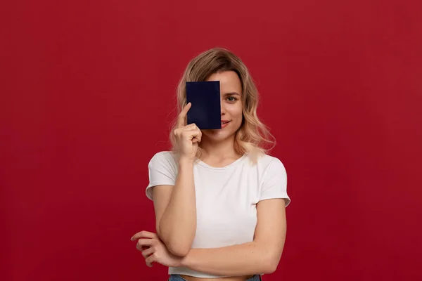 Portrait d'une fille aux cheveux blonds bouclés dans un t-shirt blanc debout sur un fond rouge. Modèle sourit à la caméra et détient un passeport de couleur bleue couvrant la moitié d'un visage . — Photo