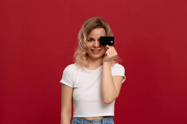 Retrato de una chica con el pelo rubio rizado en una camiseta blanca de pie sobre un fondo rojo. Modelo sonríe a la cámara y sostiene la tarjeta bancaria que cubre la mitad de una cara . —  Fotos de Stock