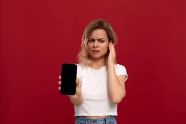 Kırmızı arka plan üzerinde beyaz bir t-shirt kıvırcık sarı saçlı bir kızın portresi. Kablosuz kulaklıktaki üzgün model kameraya bakar ve cep telefonunun ekranını gösterir.
