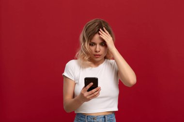 Kırmızı arka plan üzerinde beyaz bir t-shirt kıvırcık sarı saçlı bir kızın portresi. Kablosuz kulaklıkta memnun olmayan görünüme sahip model cep telefonunun ekranına bakıyor.