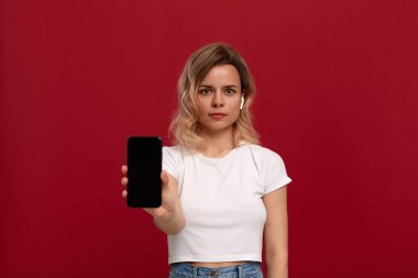 Kırmızı arka plan üzerinde beyaz bir t-shirt kıvırcık sarı saçlı bir kızın portresi. Kablosuz kulaklıktaki model kameraya bakar ve cep telefonunun ekranını gösterir. 