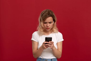 Kırmızı arka plan üzerinde beyaz bir t-shirt kıvırcık sarı saçlı bir kızın portresi. Kablosuz kulaklıkta memnun olmayan görünüme sahip model cep telefonunun ekranına bakıyor. 