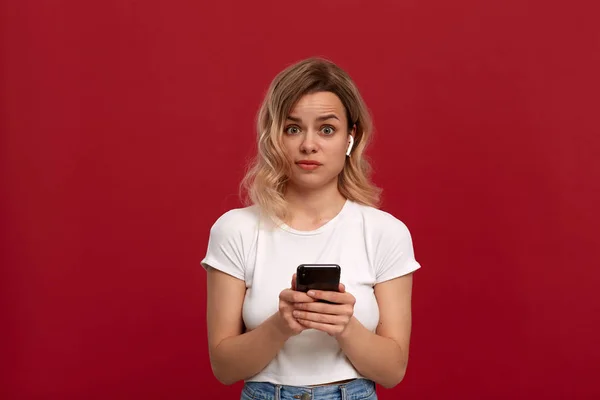 Portrait d'une fille aux cheveux blonds bouclés dans un t-shirt blanc sur fond rouge. Modèle insatisfait dans le casque sans fil regarde la caméra et tient le téléphone mobile . — Photo