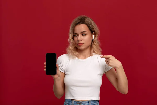 Portrait d'une fille aux cheveux blonds bouclés dans un t-shirt blanc sur fond rouge. Modèle surpris dans le casque sans fil regarde la caméra et montre l'écran d'un téléphone mobile . — Photo