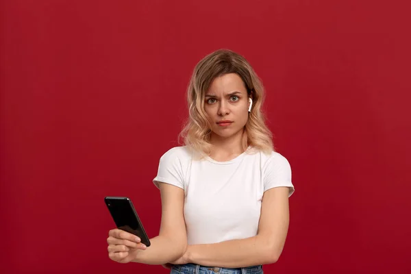 Retrato de una chica con el pelo rubio rizado en una camiseta blanca sobre un fondo rojo. Modelo con mirada insatisfecha en auriculares inalámbricos mira a la cámara y sostiene el teléfono móvil . —  Fotos de Stock