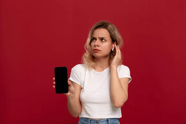 Portrait d'une fille aux cheveux blonds bouclés dans un t-shirt blanc sur fond rouge. Modèle surpris avec regard coûteux dans le casque sans fil montre l'écran d'un téléphone mobile . — Photo