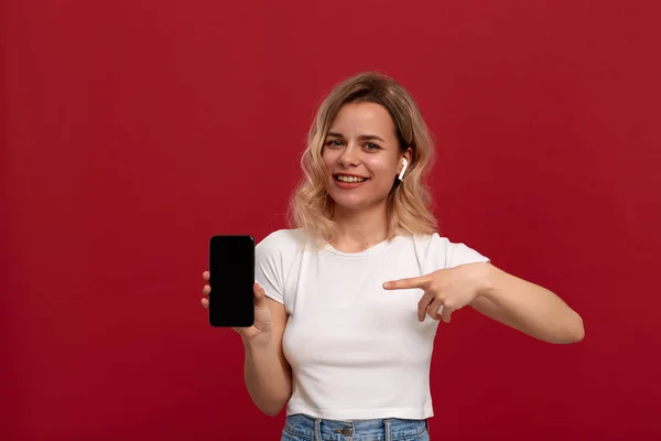 Portrait d'une fille aux cheveux blonds bouclés dans un t-shirt blanc sur fond rouge. Modèle heureux dans casque sans fil regarde la caméra et montre l'écran d'un téléphone mobile avec un doigt . — Photo