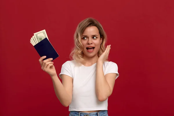 Porträtt av en flicka med lockigt blont hår klädd i en vit t-shirt stående på en röd bakgrund. Upphetsad modell som innehar ett pass av blå färg med dollar. — Stockfoto