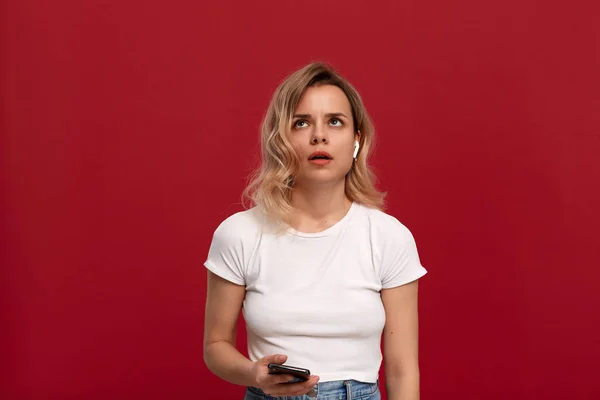 Portrait of a girl with curly blond hair in a white t-shirt on a red background. Bored model in wireless headset at lost holding mobile phone. — Stock Photo, Image