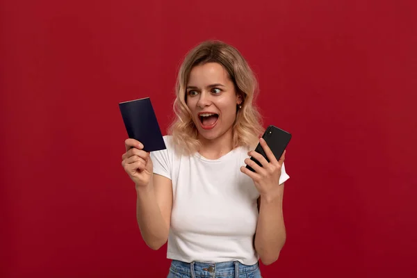 Πορτρέτο ενός κοριτσιού με σγουρά ξανθά μαλλιά σε ένα λευκό μπλουζάκι σε κόκκινο φόντο. Χαρούμενο μοντέλο κοιτάζει την οθόνη ενός κινητού τηλεφώνου και διατηρεί το διαβατήριο. — Φωτογραφία Αρχείου