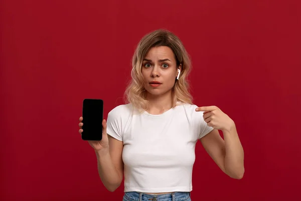 Portrait d'une fille aux cheveux blonds bouclés dans un t-shirt blanc sur fond rouge. Modèle choqué dans un casque sans fil regarde la caméra et montre l'écran d'un téléphone mobile . — Photo