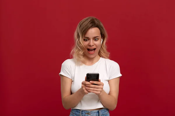 Portrait d'une fille aux cheveux blonds bouclés dans un t-shirt blanc sur fond rouge. Modèle heureux regarde l'écran d'un téléphone mobile . — Photo