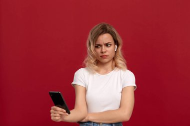 Kırmızı arka plan üzerinde beyaz bir t-shirt kıvırcık sarı saçlı bir kızın portresi. Kablosuz kulaklıkta memnun olmayan görünüme sahip model cep telefonunun ekranına bakıyor. 