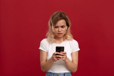 Kırmızı arka plan üzerinde beyaz bir t-shirt kıvırcık sarı saçlı bir kızın portresi. Kablosuz kulaklıkta memnun olmayan görünüme sahip model cep telefonunun ekranına bakıyor. 