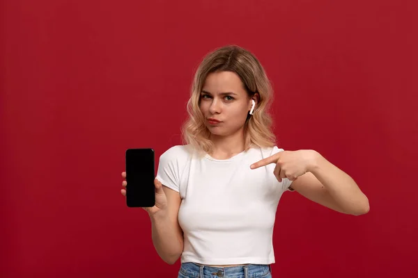 Portrait d'une fille aux cheveux blonds bouclés dans un t-shirt blanc sur fond rouge. Modèle surpris avec look incrédule dans le casque sans fil montre l'écran d'un téléphone mobile . — Photo