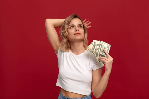 Kırmızı bir arka plan üzerinde duran beyaz bir t-shirt giymiş kıvırcık sarı saçlı bir kızın portresi. Rüya gibi bir görünüm ile dolar bir demet ile Mutlu model hayranları kendini.