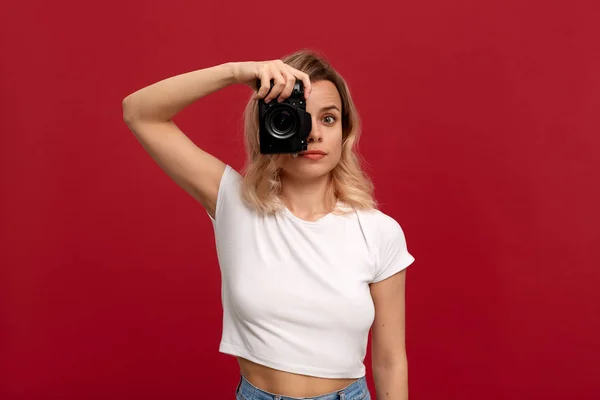 赤い背景に立つ白いTシャツに身を包んだ巻き毛のブロンドの髪の少女の肖像画。モデルはレトロなカメラで写真を撮ります. — ストック写真