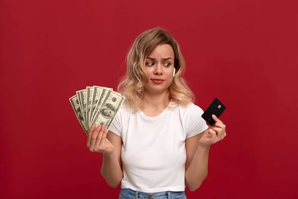 Portrait of a girl with curly blond hair in a white t-shirt standing on a red background. Model with wireless headphones looks at the bank card holding bundle of dollars expressing concern — Stock Photo, Image