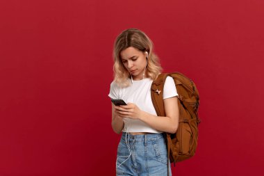 Kırmızı bir arka planda beyaz bir t-shirt ayakta giyinmiş kıvırcık sarı saçlı bir kızın portresi. Telefonda turuncu sırt çantası ve beyaz kulaklık manifatura modeli.