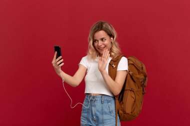 Turuncu sırt çantası ve kırmızı arka planda ayakta iken cep telefonu ile sohbet beyaz kulaklık video ile beyaz bir t-shirt giymiş kıvırcık sarı saçlı heyecanlı model.
