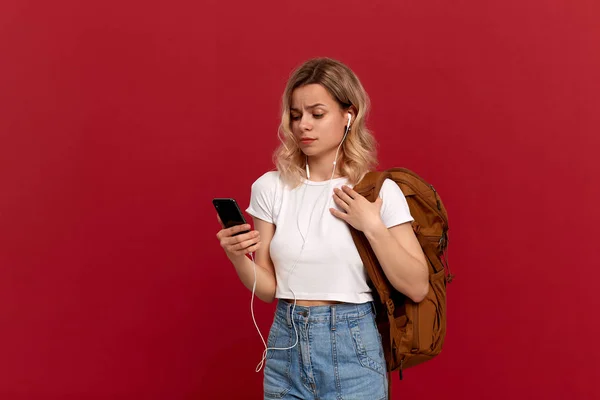 Portrait d'une fille aux cheveux blonds bouclés habillée d'un t-shirt blanc debout sur un fond rouge. Modèle avec sac à dos orange et casque blanc regarde avec scepticisme l'écran du téléphone . — Photo