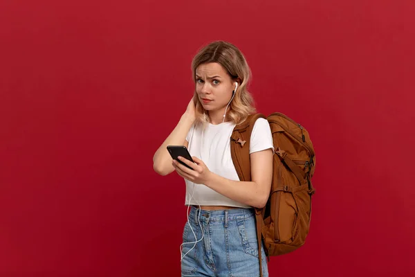 Portrait d'une fille aux cheveux blonds bouclés habillée d'un t-shirt blanc debout sur un fond rouge. Modèle choqué avec sac à dos orange, casque blanc et téléphone regarde la caméra . — Photo