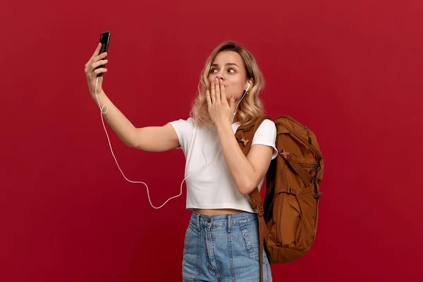 Portrait d'une fille aux cheveux blonds bouclés habillée d'un t-shirt blanc debout sur un fond rouge. Modèle excité avec sac à dos orange et casque blanc regarde l'écran du téléphone . — Photo